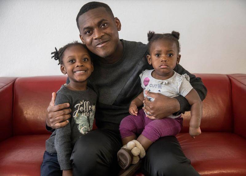 A Black man smiles big while holding his two daughters 